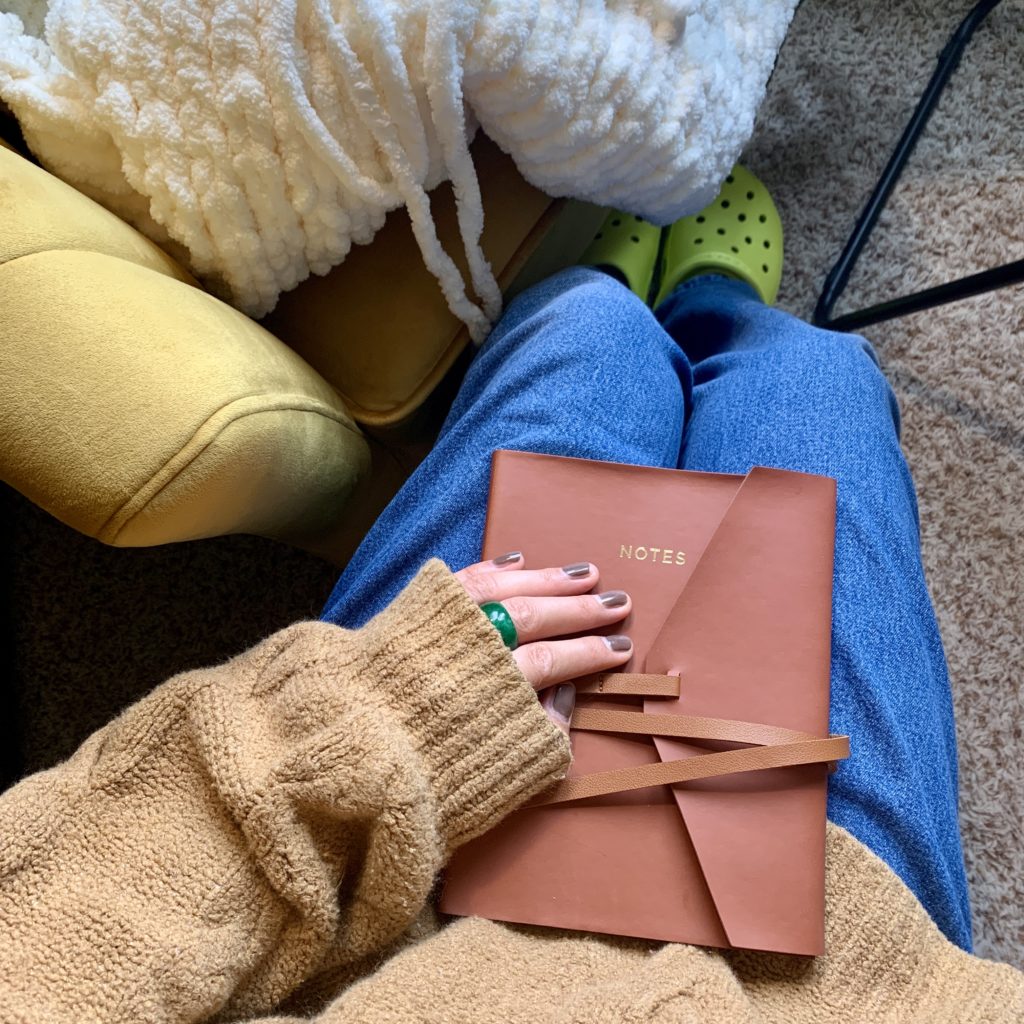 Hand on brown notebook on jeans with green clogs peeping out