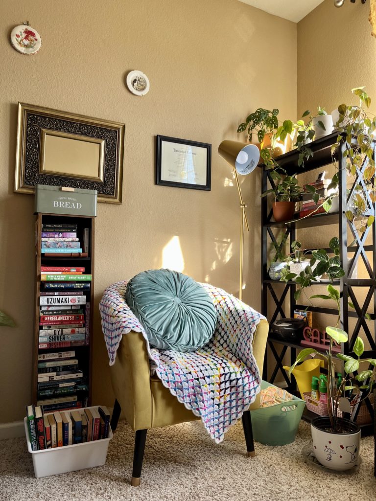 Corner of a room with a vertical shelf, plant shelf, and yellow chair with blanket in the middle